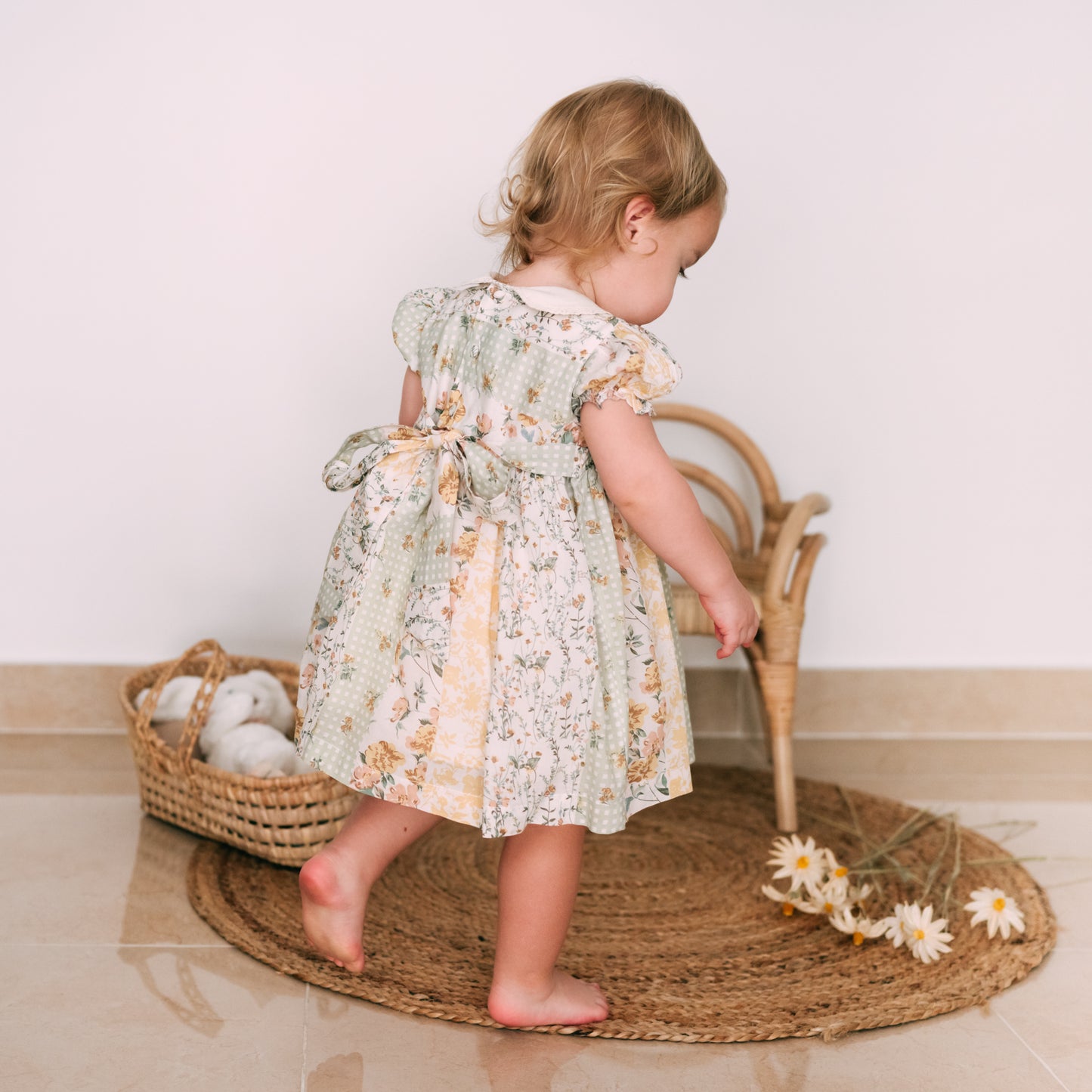 Toddler dressed in a vintage-inspired smocked floral dress, accessorized with a bow, playing near woven decor.