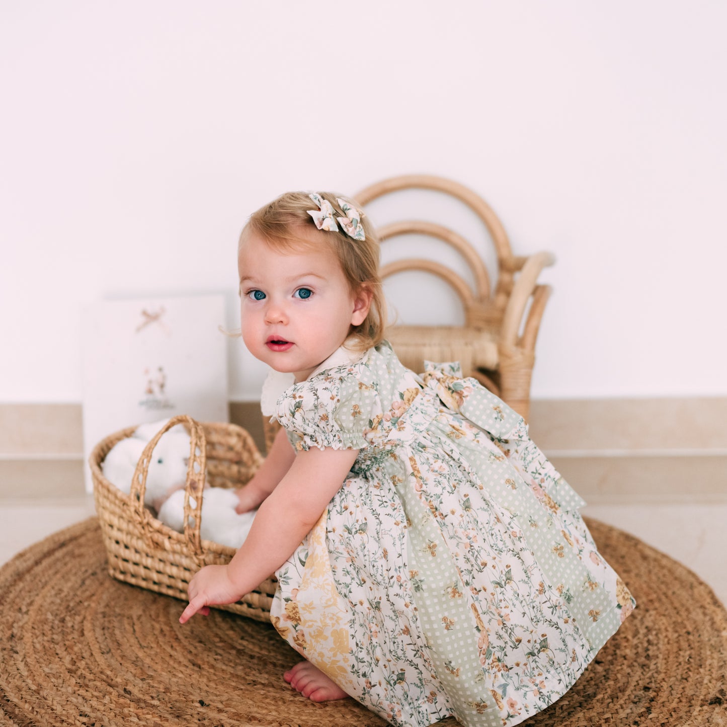 Little girl in a soft pastel dress with delicate embroidery and floral patterns, sitting by a basket of toys.