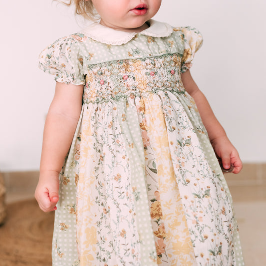 Baby girl wearing a pastel floral dress with hand-smocked embroidery, puff sleeves, and a Peter Pan collar, paired with matching accessories.