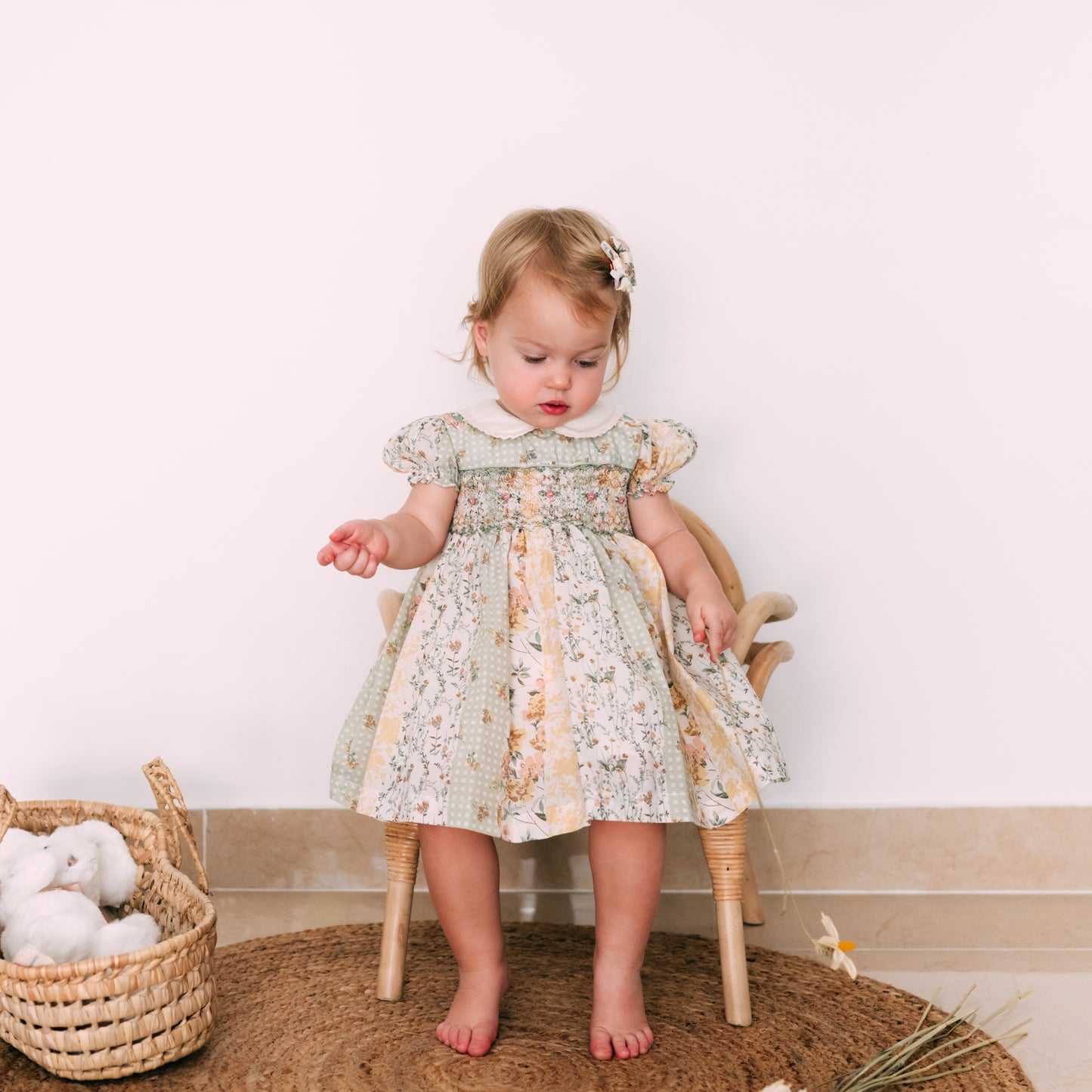 Little girl in a soft pastel dress with delicate embroidery and floral patterns, sitting by a basket of toys.