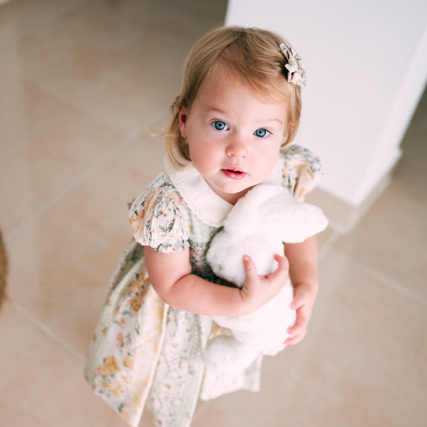 Baby girl in a smocked floral dress with a Peter Pan collar, holding a soft white bunny plush and looking up with bright blue eyes.